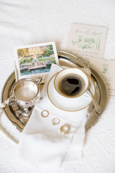 Wedding rings flatlay with engagement ring and band on a silver tray beside a coffee cup, pearls, and invitation on white bedding