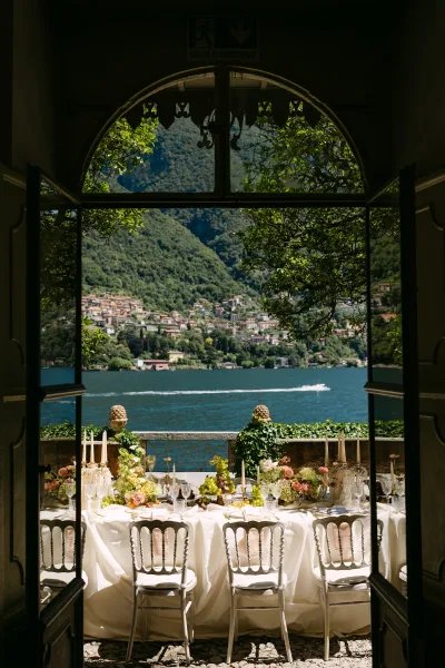 Reception tablescape for an outdoor wedding reception: long white-linen banquet table with taper candles, pastel florals, grapes, ivy, lake and mountains beyond.