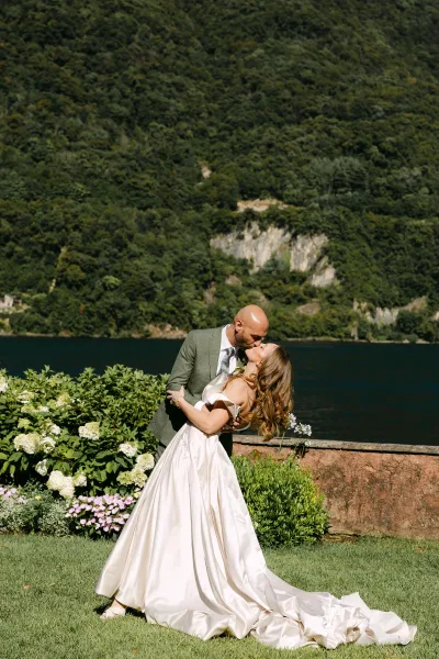 Wedding kiss portrait of groom dipping the bride in a satin ball gown, bouquet and hydrangeas by a lake with forested mountains behind