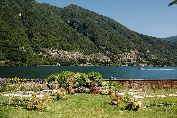 Ceremony setup for an outdoor wedding ceremony with chair rows, floral aisle border, and arch on a lawn by a lake and mountains