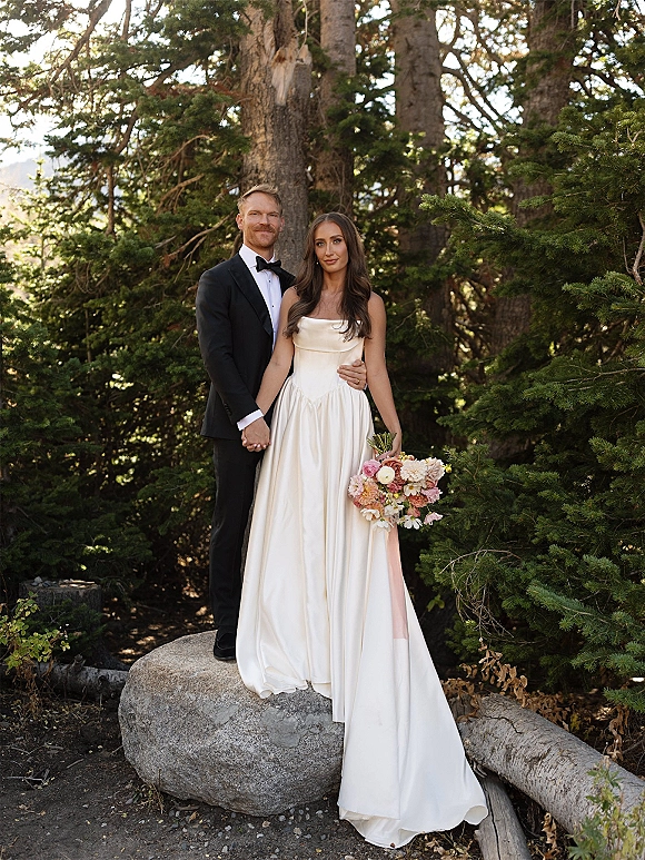 Couple portrait of bride and groom holding hands, bride in satin strapless gown with pastel bouquet, in an evergreen forest by a rock