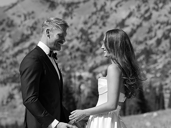 Couple portrait of bride in a strapless wedding dress and groom in tuxedo with bow tie, laughing hand in hand with mountains behind