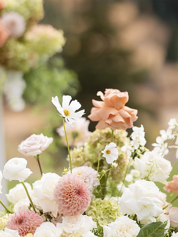 Wedding floral arrangement of pastel wedding flowers with white cosmos, blush dahlia, roses, hydrangea, and greenery in a blurred garden backdrop