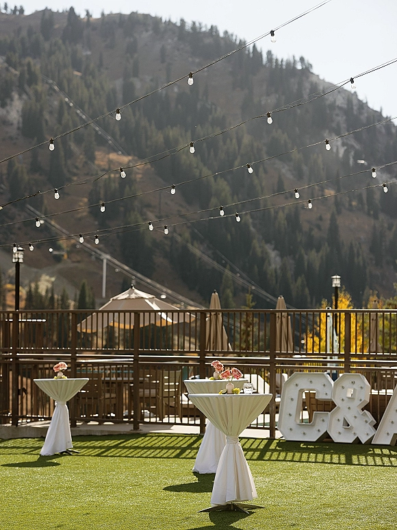 Wedding cocktail hour with white-linen high top tables, floral centerpieces and glasses under string lights on a mountain patio deck