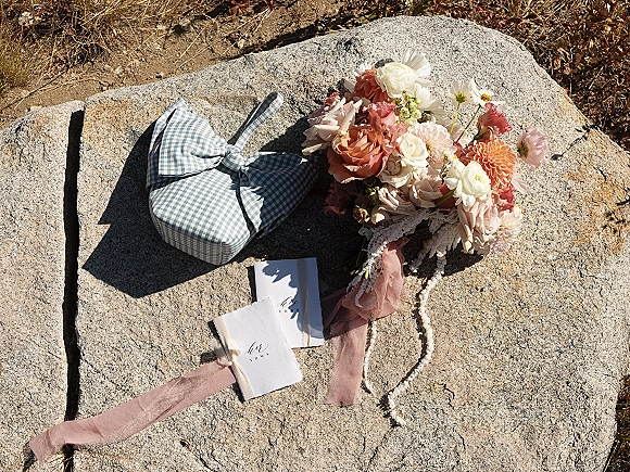 Wedding details flat lay with wedding vow books beside a peach and blush bouquet and gingham box on stone and dry grass, with pink ribbon accent