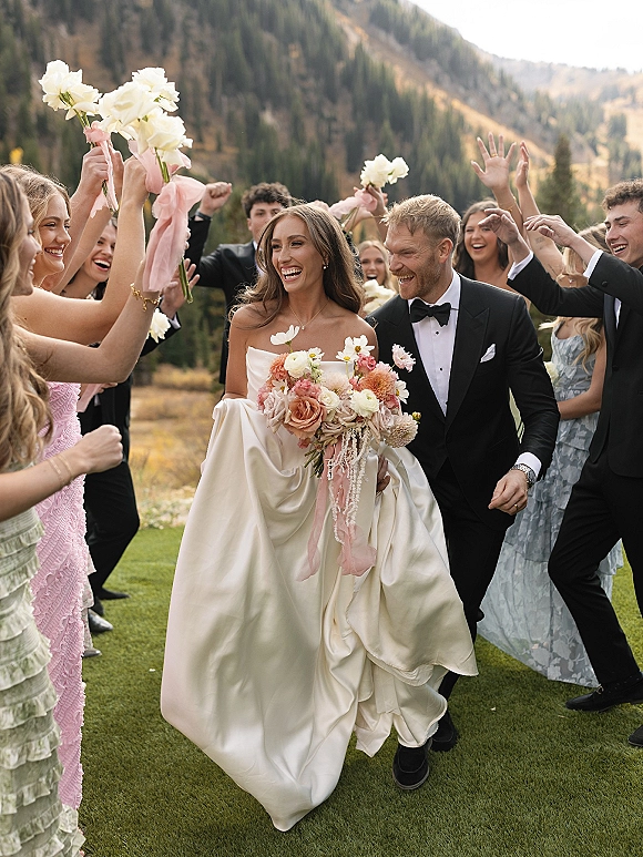 Wedding recessional as bride and groom walk the aisle smiling, bouquet ribbons flowing, with cheering guests in a mountain meadow backdrop