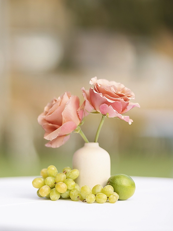 Wedding table centerpiece with a rose bud vase centerpiece holding pink roses, surrounded by green grapes and a green apple on white tablecloth