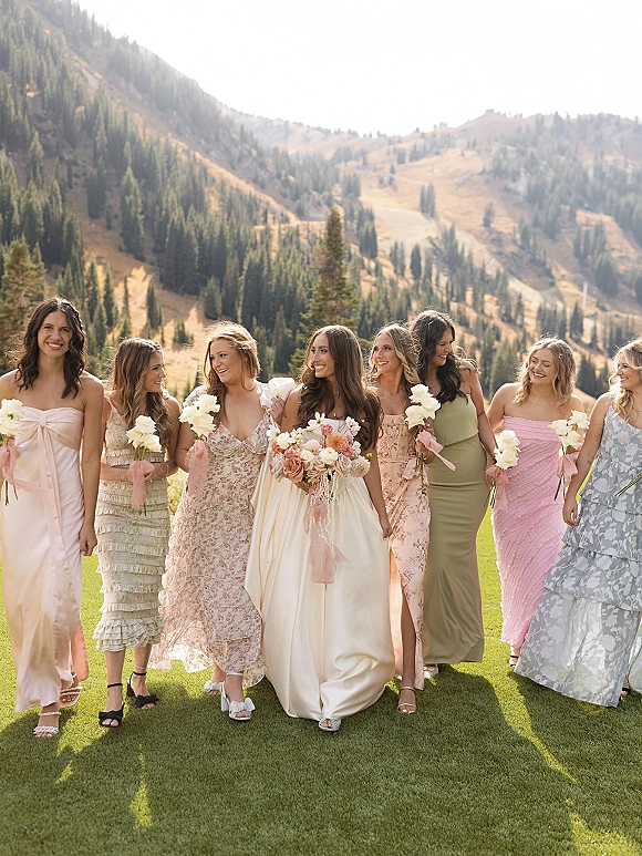 Bridesmaid group photo of bride with bridesmaids walking with mixed flower bouquets and ribbons on a grassy lawn with mountain pines behind