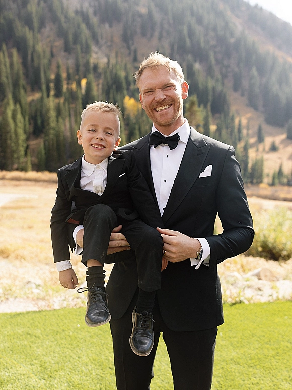 Groom with child in a black tuxedo and bow tie holds his son in a suit, posing in a sunlit grassy field with mountains and evergreens behind