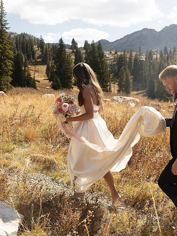 Bride portrait holding a pink and white bouquet with ribbon, strapless dress train in a mountain meadow with pines and rocks behind
