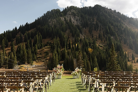 Ceremony setup with a green aisle runner lined by wood crossback chairs and pastel florals on a grassy lawn with mountains and ski lift beyond