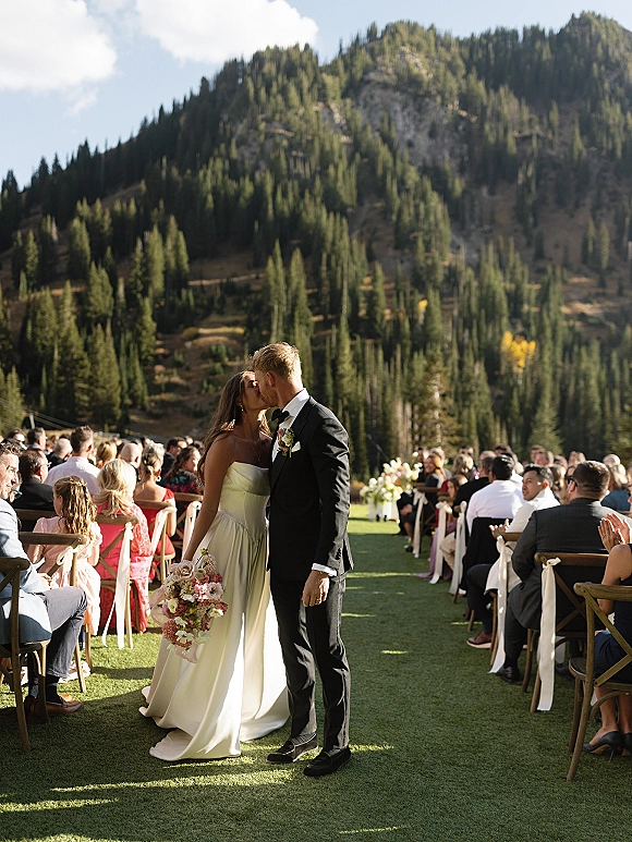 Wedding kiss as bride holds a colorful bouquet and groom in tuxedo walks the aisle, with mountain evergreens and seated guests behind