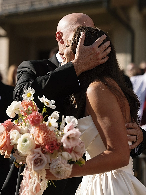 Ceremony hug as bride and groom embrace, her strapless dress and pastel daisy bouquet glowing in sunlight before an outdoor crowd
