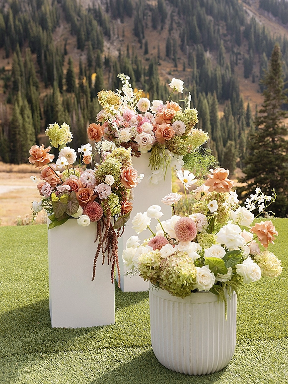 Ceremony floral arrangements on white pedestals with roses, dahlias, hydrangea, and greenery on a grassy lawn with mountain forest backdrop