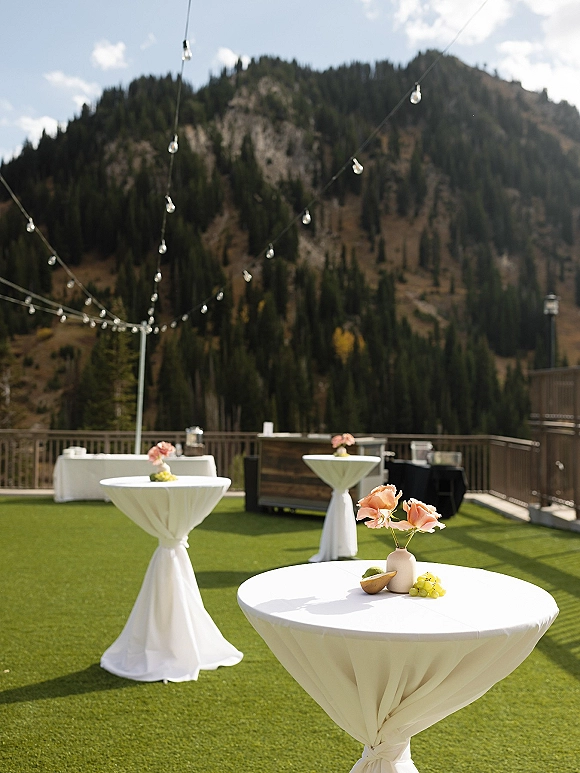 Cocktail hour setup with wedding cocktail tables in white linens, bud vase with peach flowers and grapes, under string lights on a mountain terrace