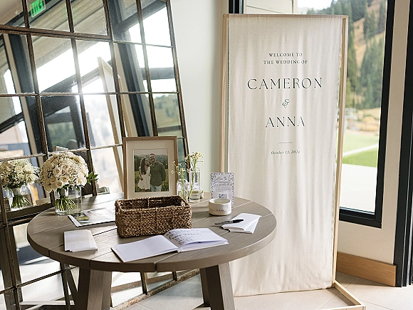 Wedding welcome sign on a round entry table with guest book, pen, framed photo, white roses and baby's breath, by windows with mountain view