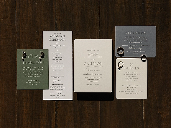 Invitation suite flat lay with wedding rings and pearl drop earrings on printed cards, arranged on a dark wood tabletop