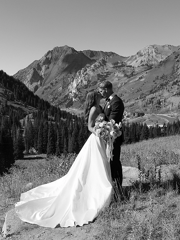 Wedding kiss portrait of bride holding a bouquet and groom in a tuxedo, kissing in a hillside meadow with pine forest and mountains behind