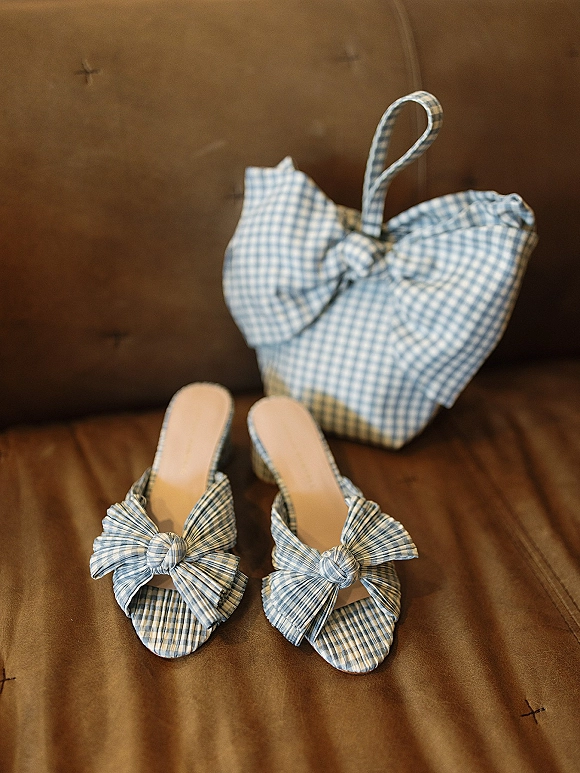 Bridal shoes in blue gingham with bow heels beside a matching gingham handbag on a tufted brown leather sofa