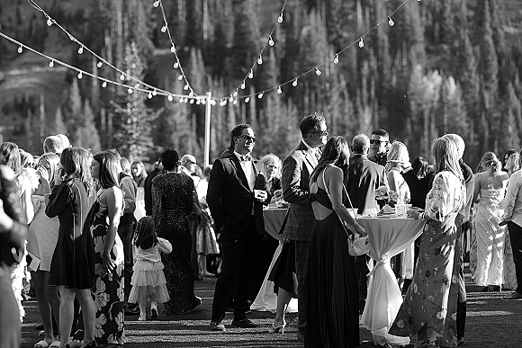 Wedding cocktail hour with outdoor cocktail hour guests mingling around high-top tables under string lights on a mountain lawn by pines