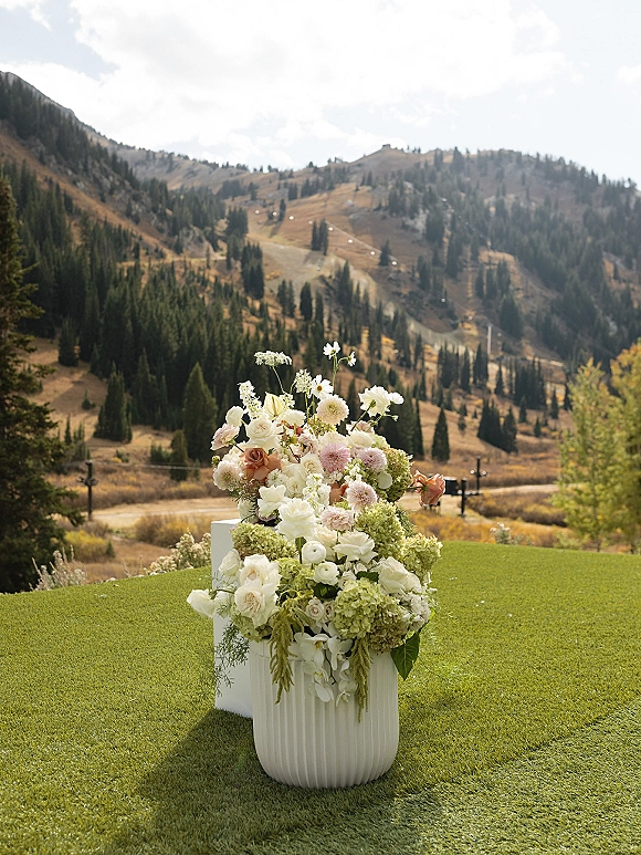 Wedding floral arrangement on a white pedestal plinth with roses, dahlias, hydrangea, and hanging greenery against mountain scenery