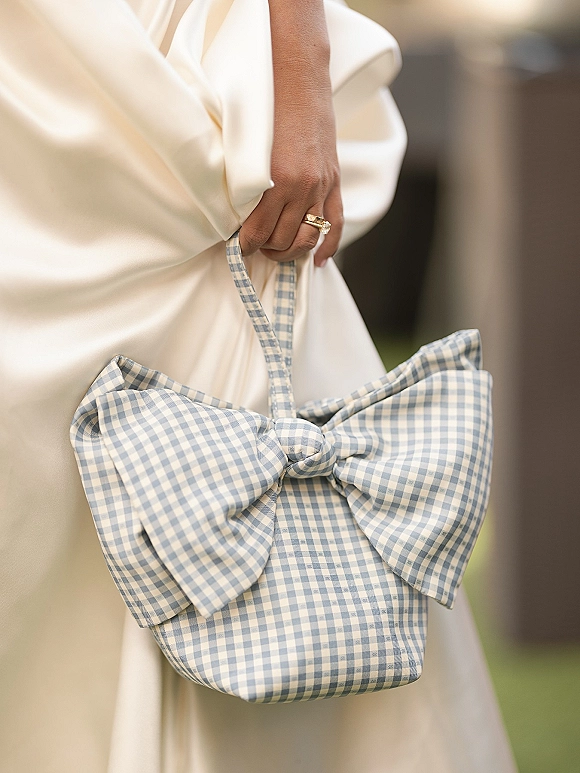 Bridal purse, wedding day handbag with a blue gingham bow held against a satin gown, showing engagement ring and band on an outdoor lawn