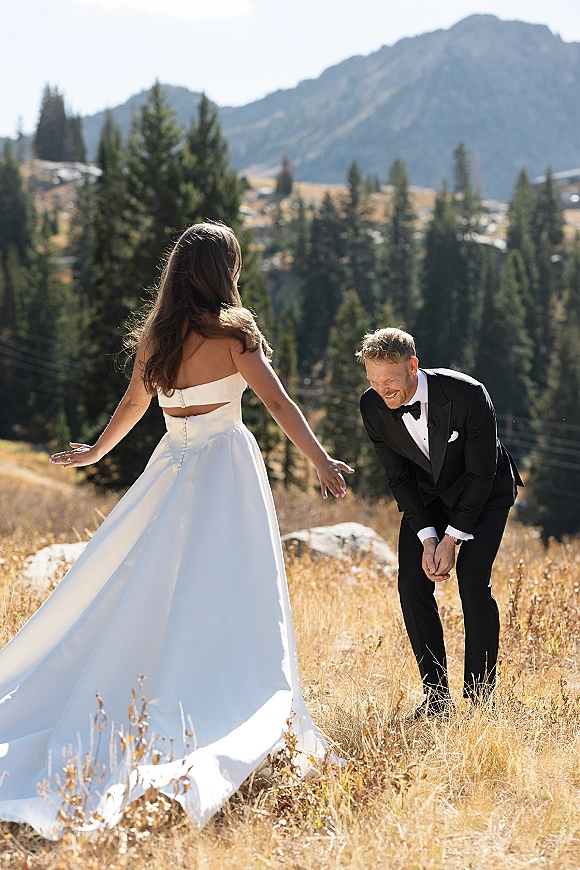 Wedding first look as bride in a strapless gown approaches groom in black tuxedo with bow tie, laughing in a mountain meadow with pines