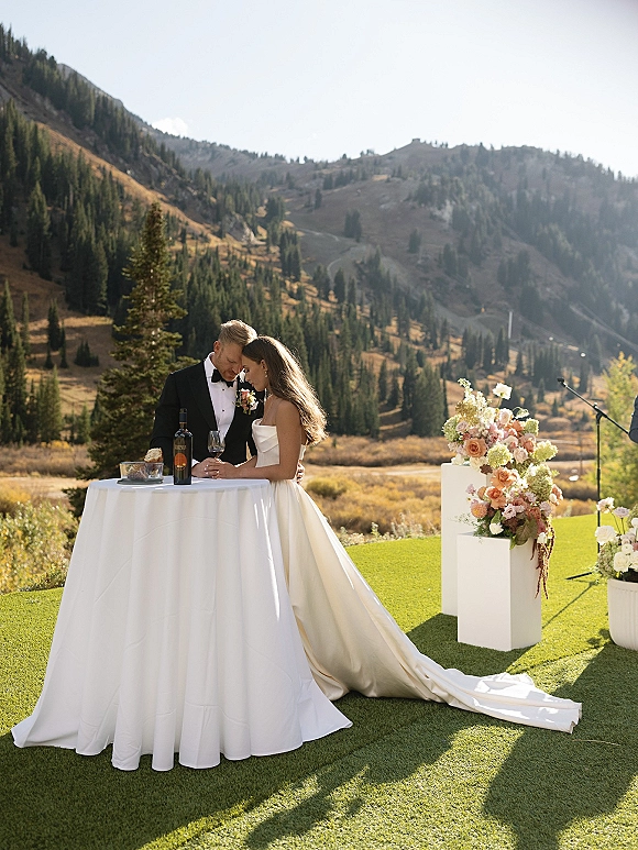 Couple portrait of bride in strapless gown with long train and groom in tuxedo beside a cocktail table, mountains and pines behind