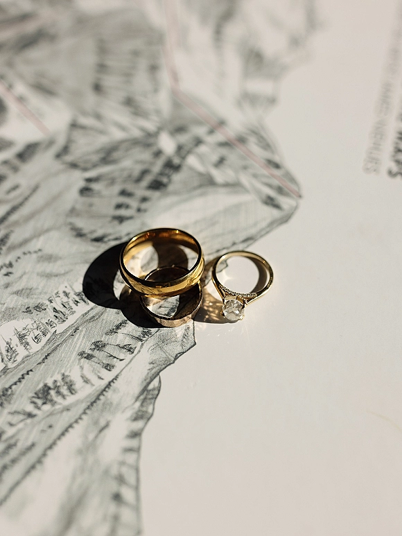 Wedding rings with a gold wedding band and diamond engagement ring stacked on white illustrated paper, casting crisp hard sunlight shadows
