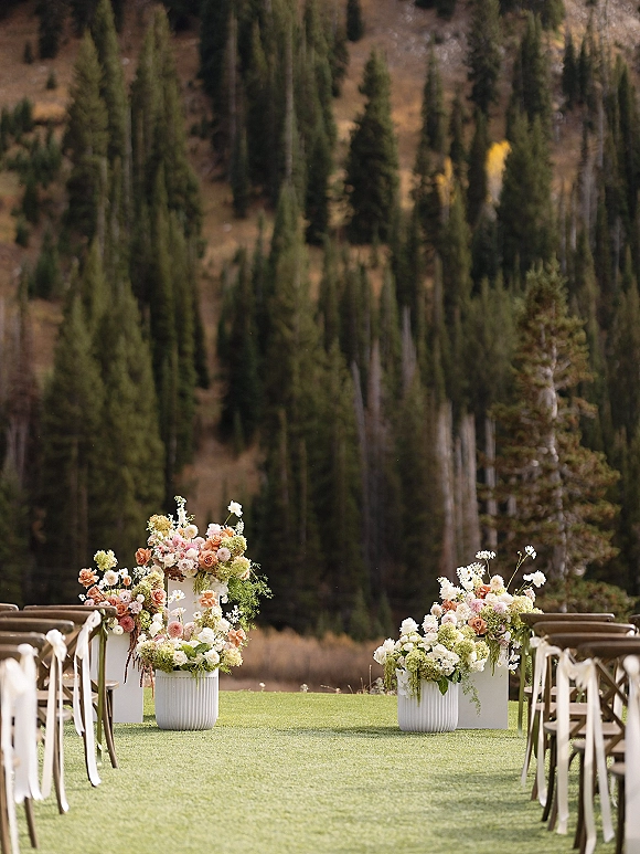 Ceremony aisle decor with floral arrangements on white pedestal planters and ribboned chairs lining a grassy path in a pine forest clearing