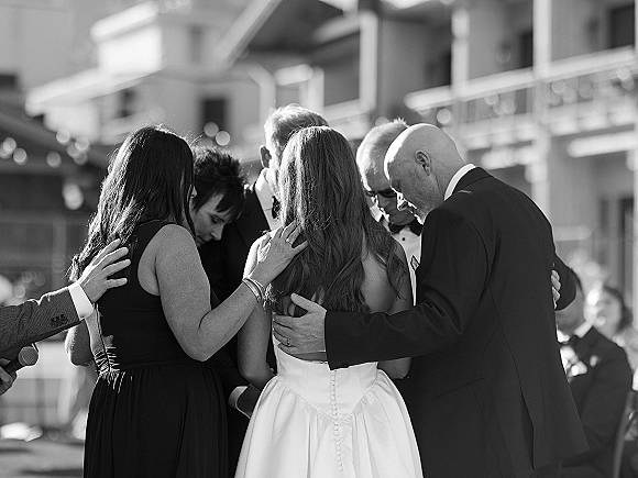 Wedding prayer with bridal party in a ceremony prayer circle, hands on shoulders around the bride as guests watch from a balcony railing