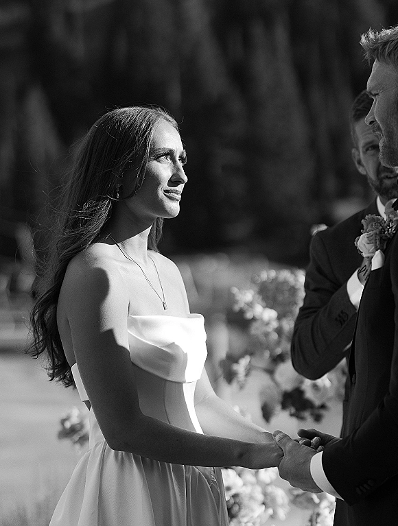 Wedding vows shared as the couple holds hands, bride in a strapless dress with necklace and drop earrings at an outdoor ceremony with trees behind
