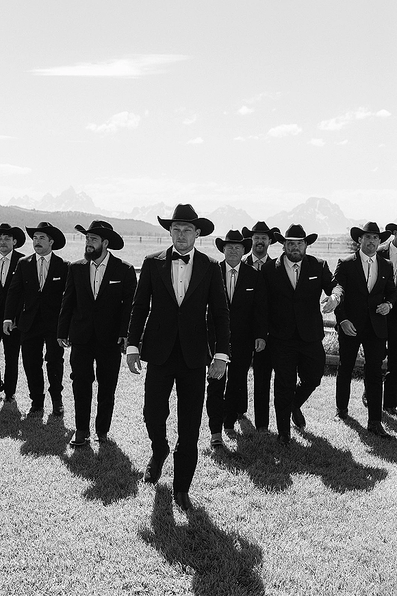Groomsmen portrait with cowboy hats and tuxedos walking across a grassy field, mountains and dramatic clouds behind them