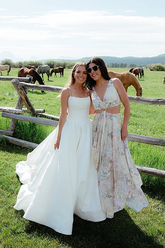 Bride with bridesmaid portrait as the bride in a strapless wedding dress hugs her friend by a wooden fence with horses and mountains behind