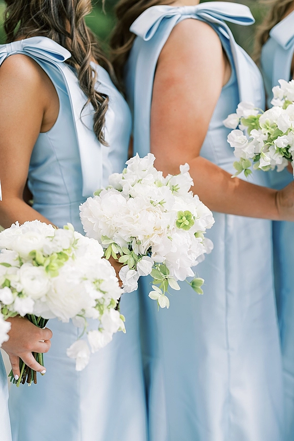 Bridesmaid dresses in light blue bridesmaid dresses with shoulder bows, holding white bouquets with greenery against blurred outdoor foliage