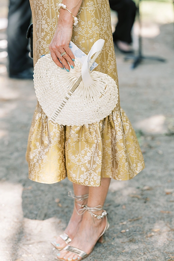 Wedding guest outfit featuring a gold midi wedding guest dress with woven handbag, pearl bracelet, metallic heels, on outdoor pavement by blurred guests