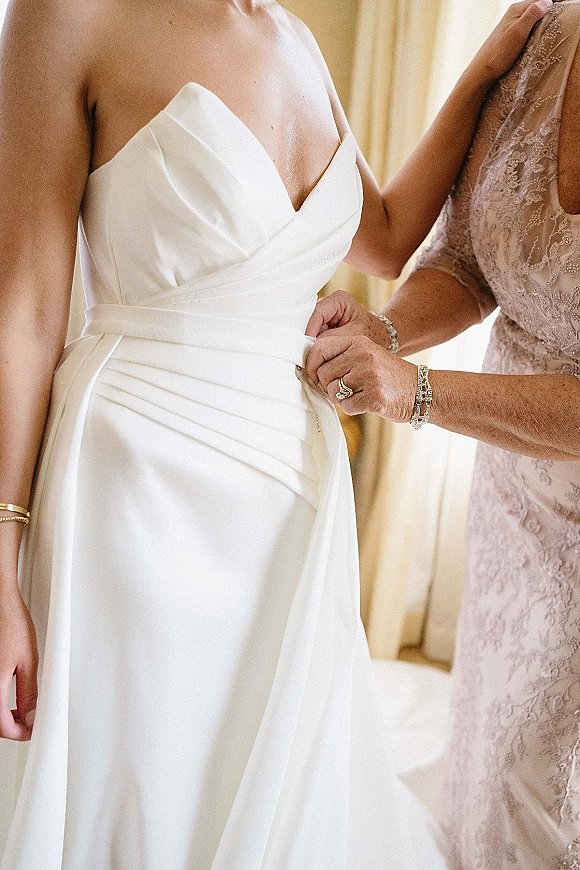 Wedding dress close-up of a strapless wedding dress with satin waist pleats, showing the bride’s ring and bracelet near soft curtains indoors