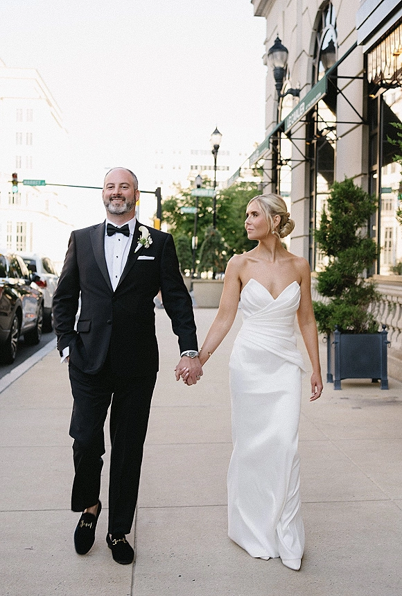 Couple portrait of bride and groom walking hand in hand on a city sidewalk, her strapless gown and his black tuxedo with boutonniere