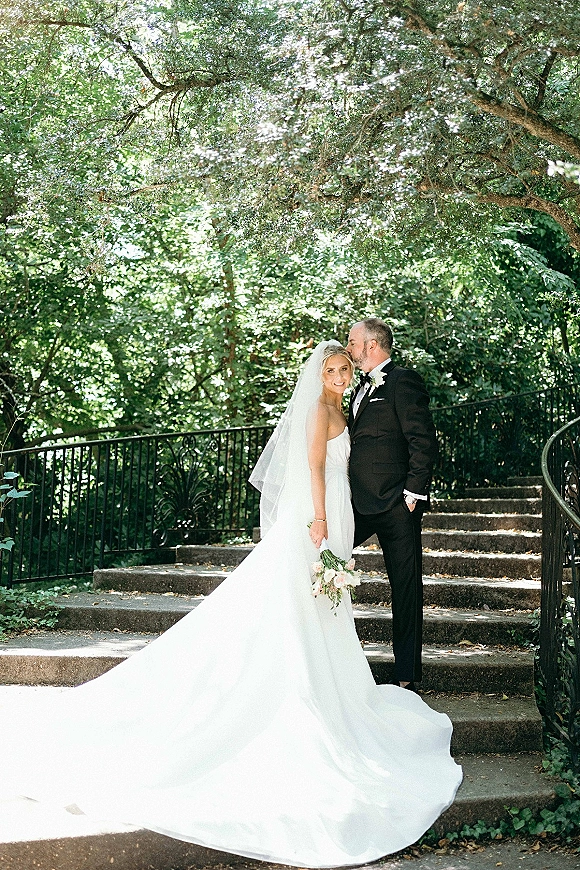 Couple portrait of groom kissing bride’s forehead on a stone staircase, her veil and long train flowing as she holds a bouquet