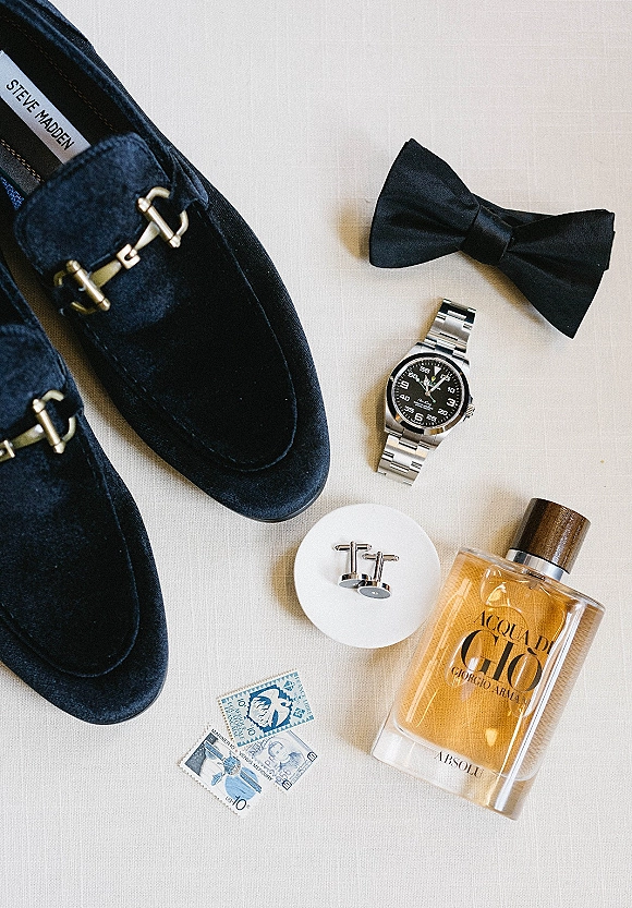 Groom accessories flatlay with black velvet loafers, black bow tie, silver wristwatch, cufflinks, cologne, and postage stamps on white fabric