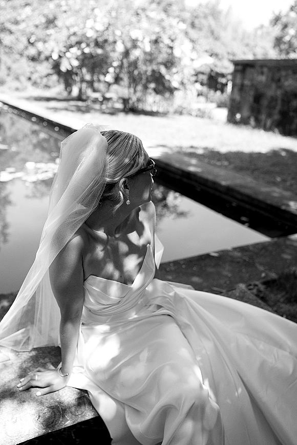 Bridal portrait of a bride in sunglasses and strapless dress with long veil, seated by a reflecting pool on a stone ledge in a garden