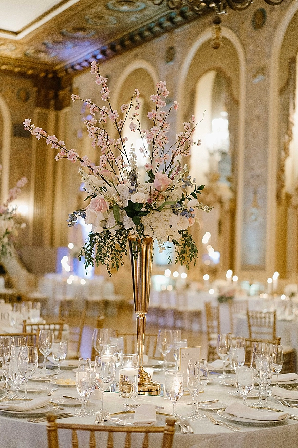 Reception tablescape with tall wedding centerpiece in a gold trumpet vase, blush and white florals, candlelight, and ornate mirrored ballroom backdrop