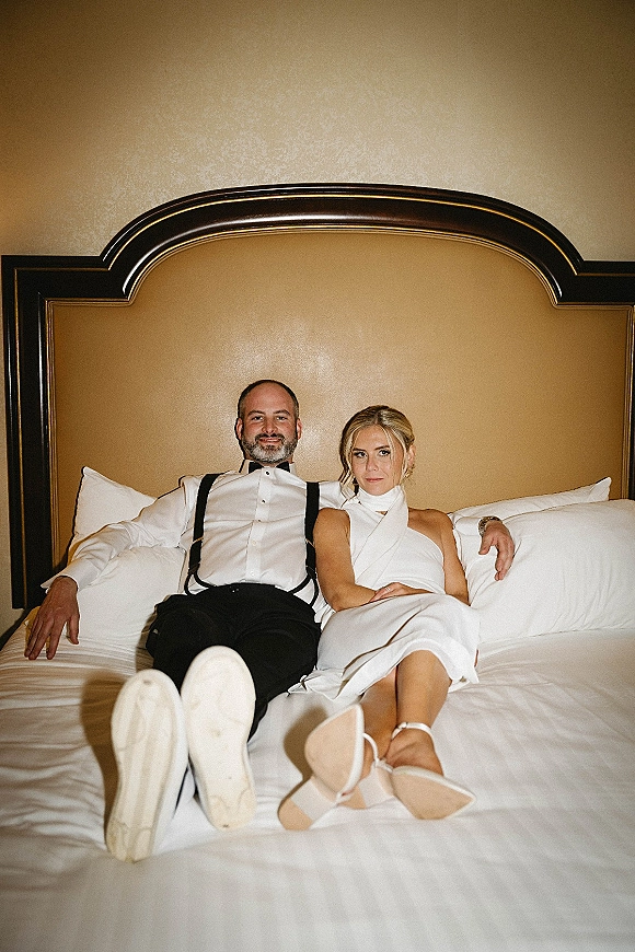 Couple portrait of newlyweds on bed in a hotel room, bride in a white halter mini dress and heels, groom in bow tie and suspenders