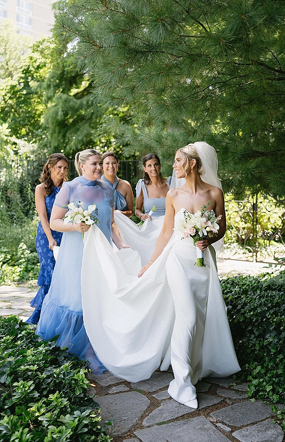 Bride with bridesmaids walking along a stone garden path, bride in strapless satin gown with veil holding a white blush bouquet