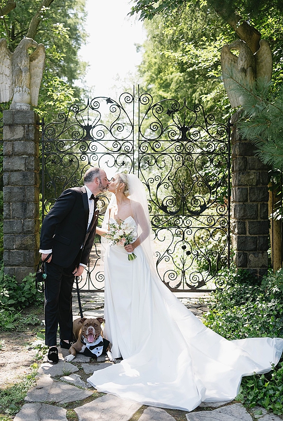 Wedding kiss portrait of bride and groom kissing in a garden, bouquet and veil in hand, with a tuxedoed dog on leash by a wrought iron gate