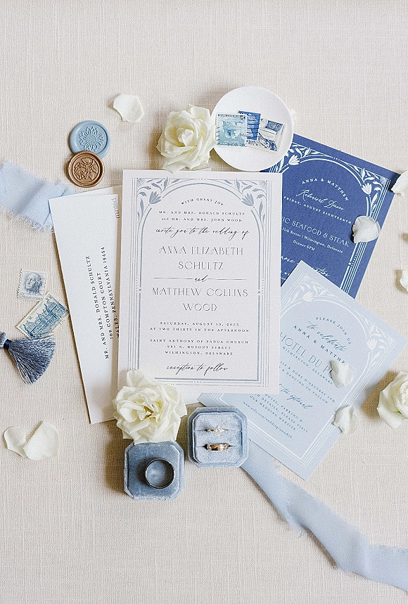 Wedding invitation suite with blue and white wedding invitations, letterpress cards, wax seal, vintage stamps, rings, and white roses on linen backdrop