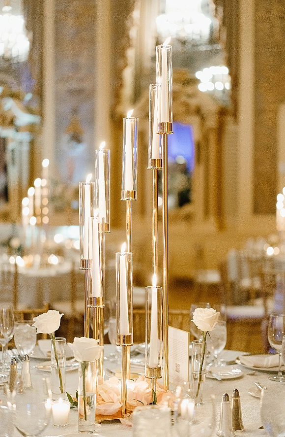 Wedding table centerpiece with tall candle centerpiece, white and blush roses and taper candles glowing on a reception table under warm lighting