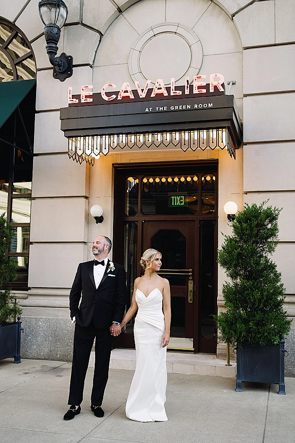 Couple portrait of bride and groom holding hands in formal attire by a stone arched venue entrance with marquee lights and neon sign.