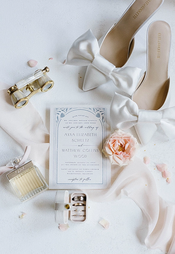 Invitation suite wedding invitation flat lay with rings in a box, white bow heels, perfume, pink rose, and opera glasses on white fabric backdrop
