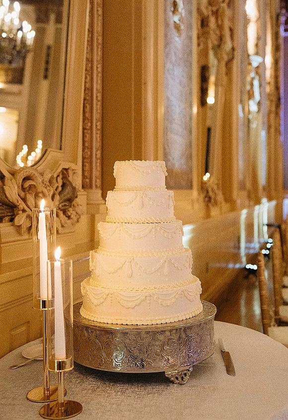 Wedding cake four tier wedding cake in white buttercream with garland piping on a silver stand, flanked by taper candles in a gilded ballroom